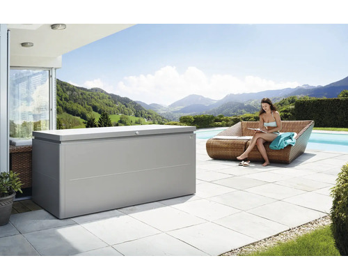 Boîte de rangement biohort LoungeBox 200, 200x84x88,5 cm gris quartz métallique Scène d''extérieur avec un grand coffre de rangement à côté d''une femme assise sur un canapé au bord d''une piscine, avec vue sur un paysage montagneux