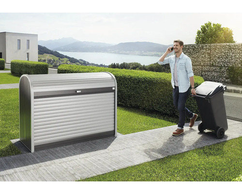 Cache-poubelle biohort StoreMax 160 163 163x78x120 cm gris foncé métallique Cache-conteneur à ordures en métal avec volet roulant dans le jardin, un homme téléphone et tire une poubelle
