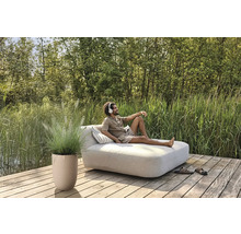 Scène avec un homme sur une chaise longue de jardin avec coussins, plante en pot et chaussures sur une terrasse en bois au bord d''un lac