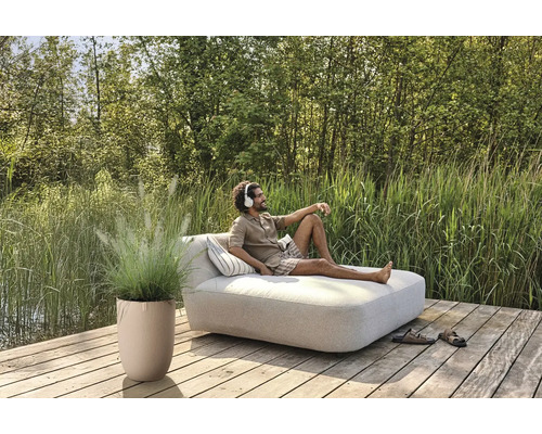 Scène avec un homme sur une chaise longue de jardin avec coussins, plante en pot et chaussures sur une terrasse en bois au bord d''un lac