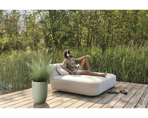 Un homme se détend avec un casque sur un transat de jardin, sur une terrasse en bois, à côté d''un bac à plantes avec des graminées ornementales.
