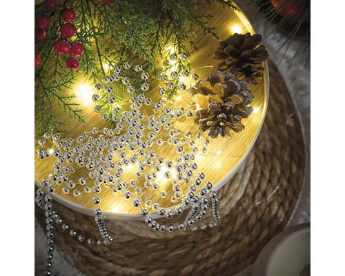 Décoration avec collier de perles argentées, branches de sapin et pommes de pin sur une table ronde.