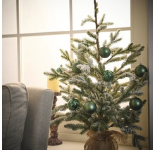 Dekorativer Tannenbaum mit grünen Kugeln und Jutesack auf einem Tisch vor einem Fenster.