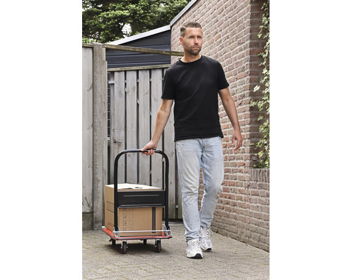 Un homme tire un chariot de manutention avec des cartons sur un chemin pavé.