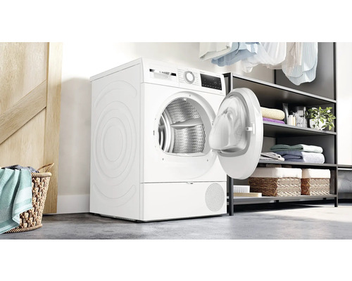 Sèche-linge blanc avec porte ouverte dans une pièce lumineuse avec des étagères et des paniers à linge.