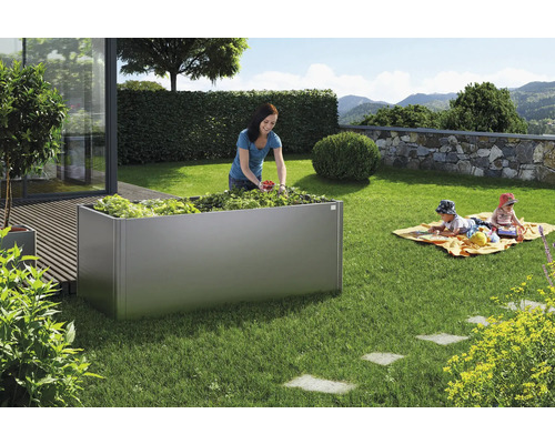 Scène de jardin avec un potager surélevé en métal, une femme récoltant des fraises et des enfants jouant à l''arrière-plan.