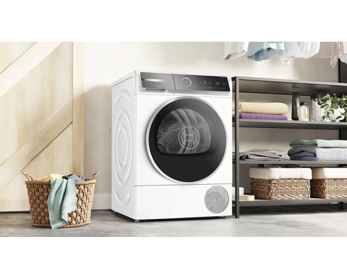 Freistehender weisser Bosch Wärmepumpentrockner neben einem Regal und einem Wäschekorb.