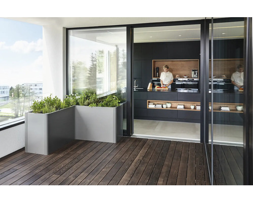 Scène avec des herbes aromatiques dans des bacs à plantes gris sur un balcon en bois devant une porte coulissante ouverte donnant sur une cuisine