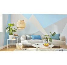 Wohnzimmer mit Sofa, Tisch, Lampe und Zimmerpflanzen vor einer Wand mit geometrischen Mustern