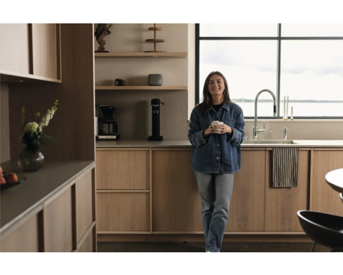 Helle Küche mit Holzfronten, Edelstahlarmatur und Fenster mit Blick auf das Wasser. Eine Frau hält eine Tasse in der Hand.