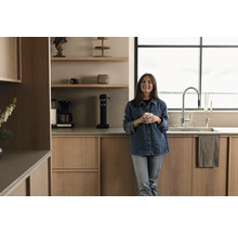 Helle Küche mit Holzschränken, Spüle, Armatur und einer Frau mit einer Tasse in der Hand.
