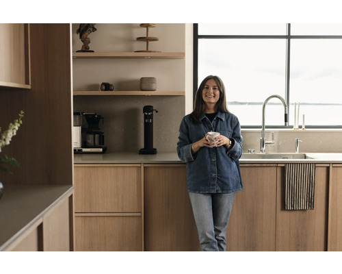Helle Küche mit Holzschränken, Spüle, Armatur und einer Frau mit einer Tasse in der Hand.