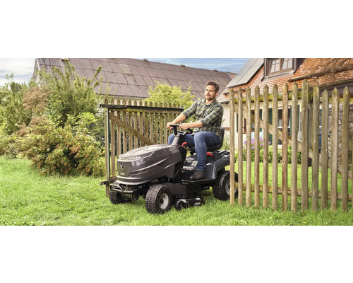 Un homme conduit un tracteur de pelouse sur une pelouse à côté d'une clôture en bois.