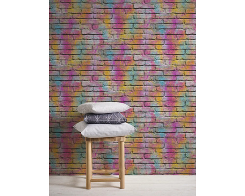 Mur avec un papier peint en briques colorées et une pile de coussins sur un tabouret.