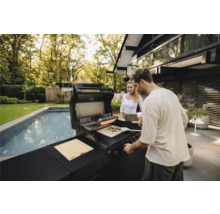 Un homme et une femme préparent une pizza sur un grill extérieur à côté d'une piscine