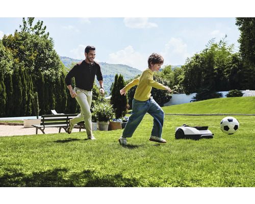 Un homme et un enfant jouent au football avec un robot tondeuse dans le jardin.