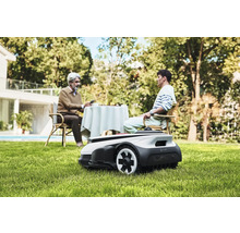 Scène avec un robot tondeuse à gazon dans le jardin, tandis que deux personnes sont assises à une table