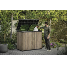 Cache-poubelle en bois avec deux poubelles vertes et une personne jetant un sac poubelle à l'intérieur