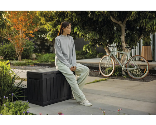 Szene im Garten mit einer sitzenden Person auf einer Aufbewahrungsbox und einem Fahrrad im Hintergrund