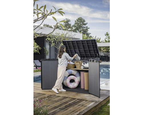 Une femme range une boîte de jardin grise contenant des accessoires de piscine sur une terrasse en bois.