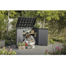 Jardin avec une femme agenouillée devant un abri de jardin ouvert dans lequel sont rangés des outils de jardinage.