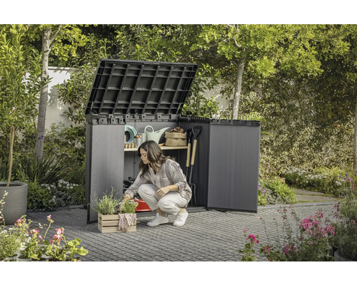 Jardin avec une femme agenouillée devant un abri de jardin ouvert dans lequel sont rangés des outils de jardinage.