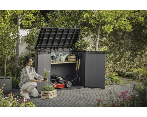 Une femme plante des fleurs devant un abri de jardin ouvert avec des outils de jardinage