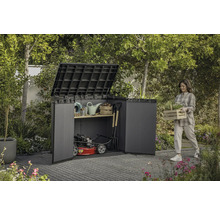 Scène de jardin avec une femme portant une caisse de plantes à côté d'un abri à outils rempli d'outils de jardin et d'une tondeuse à gazon