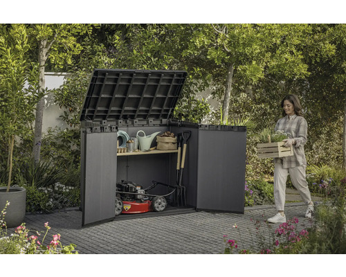 Scène de jardin avec une femme portant une caisse de plantes à côté d'un abri à outils rempli d'outils de jardin et d'une tondeuse à gazon