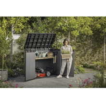 Scène de jardin avec abri de jardin, outils de jardinage et une femme tenant une caisse de plantes.