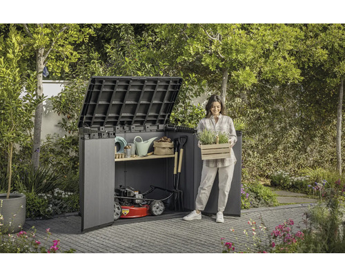 Scène de jardin avec abri de jardin, outils de jardinage et une femme tenant une caisse de plantes.