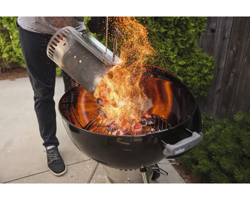 Une personne verse du charbon de bois incandescent d'une cheminée d'allumage dans un barbecue sphérique.