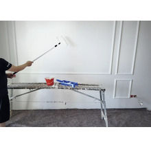 Un peintre peint un mur avec un rouleau à peinture, un seau de peinture et un bac à peinture sont posés sur un échafaudage.