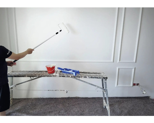 Un peintre peint un mur avec un rouleau à peinture, un seau de peinture et un bac à peinture sont posés sur un échafaudage.