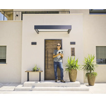 Entrée moderne avec une porte d'entrée aspect bois, un auvent sombre et des appliques extérieures. Un homme se tient devant la porte.