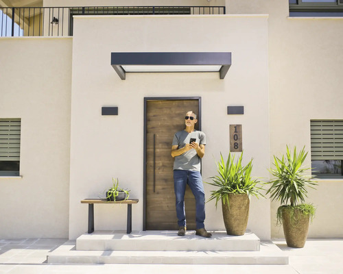 Entrée moderne avec une porte d'entrée aspect bois, un auvent sombre et des appliques extérieures. Un homme se tient devant la porte.