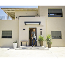 Maison moderne aux façades claires, volets roulants gris, marquise noire au-dessus de la porte d'entrée et grands bacs à fleurs dans l'entrée.