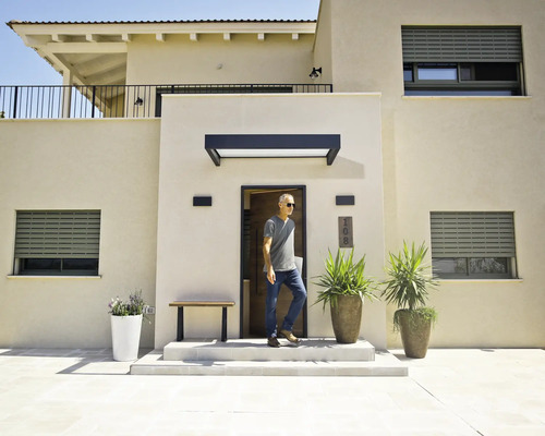 Maison moderne aux façades claires, volets roulants gris, marquise noire au-dessus de la porte d'entrée et grands bacs à fleurs dans l'entrée.