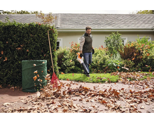 Un homme utilise un souffleur de feuilles dans le jardin, à côté d'un sac à déchets verts et d'un râteau.