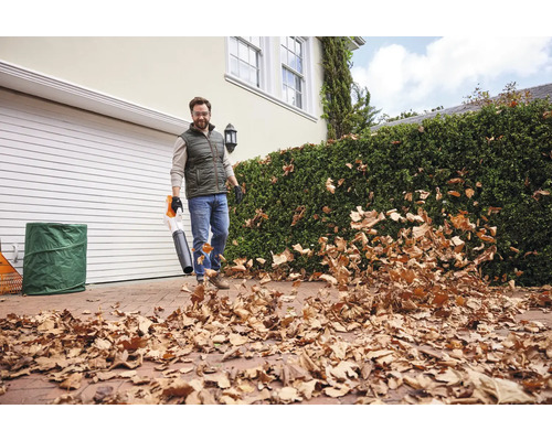 Un homme utilise un aspirateur à feuilles pour enlever les feuilles. À l'arrière-plan, on voit une maison avec un garage et une haie verte.