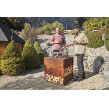 Deux hommes font des grillades sur un barbecue en acier Corten dans le jardin, rempli de bûches.