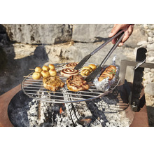 Des grillades comme de la viande, des pommes de terre et des saucisses grillées sur une grille au-dessus d'un feu ouvert, maintenues avec une pince à barbecue.