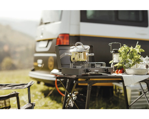 Cuisine de camping avec réchaud à gaz, casserole, lanterne et aliments devant un camping-car
