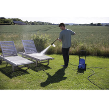Un homme nettoie deux chaises longues de jardin en bois avec un nettoyeur haute pression à l'extérieur.