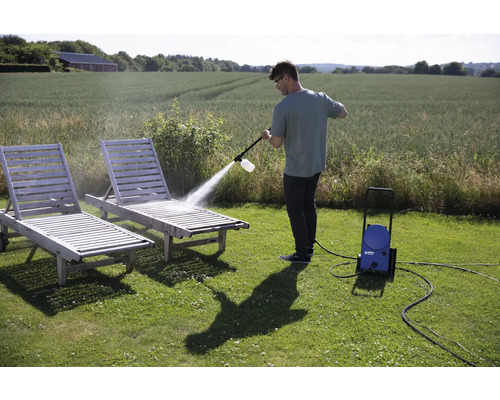 Un homme nettoie deux chaises longues de jardin en bois avec un nettoyeur haute pression à l'extérieur.