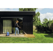 Femme nettoie une terrasse en bois avec un nettoyeur haute pression devant une maison noire