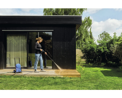 Femme nettoie une terrasse en bois avec un nettoyeur haute pression devant une maison noire