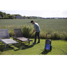 Un homme nettoie une chaise longue en bois dans le jardin avec un nettoyeur haute pression.