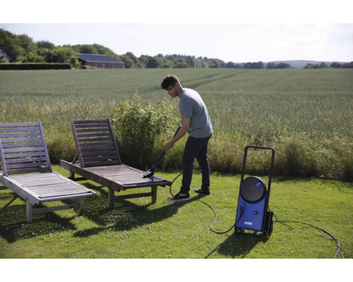Un homme nettoie une chaise longue en bois dans le jardin avec un nettoyeur haute pression.