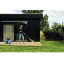 Femme nettoie terrasse en bois avec nettoyeur haute pression à côté d'une maison noire.